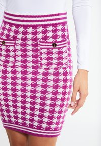 Růžová sukně se vzorem houndstooth z pleteniny s bílými akcenty, vybavená žebrovaným pasem a dvěma předními kapsami s knoflíky. Textura vypadá měkce.
