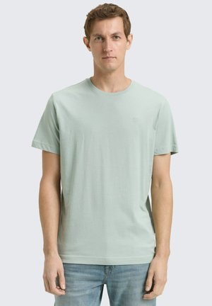 Man draagt een lichtgroen T-shirt met korte mouwen en een blauwe spijkerbroek, staand tegen een effen lichte achtergrond.