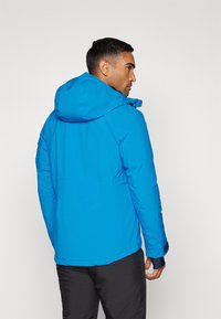 Blaue isolierte Jacke mit Kapuze, die eine glatte Textur und elastische Bündchen aufweist, von hinten gesehen, kombiniert mit schwarzen Hosen.