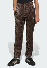 Smeđe trenirke s uzorkom tigrastih pruga, elastičnim pojasom i bočnim džepovima, uparene s crnim Adidas Superstar tenisicama.