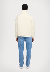 Cremefarvet, letvægtsjakke med høj krave, sænkede skuldre og elastiske manchetter, parret med lyseblå denimjeans og hvide sneakers.