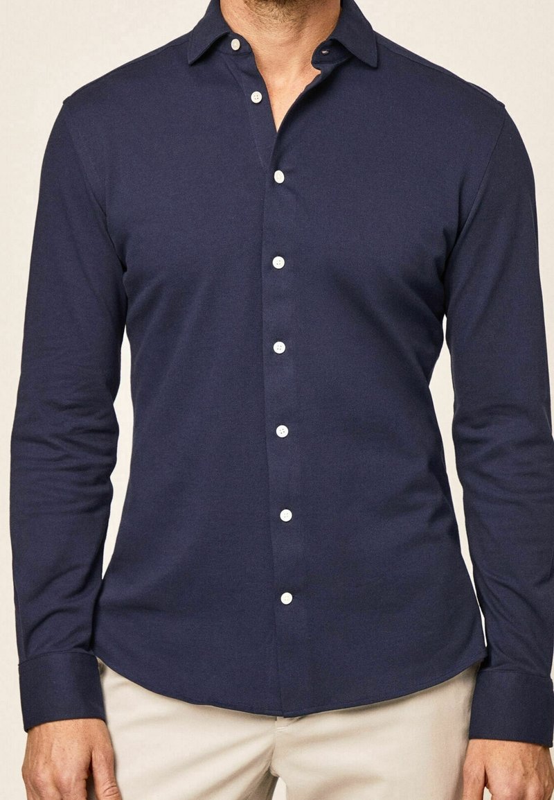 Homme portant une chemise ajustée bleu marine à manches longues avec des boutons blancs et un pantalon beige.