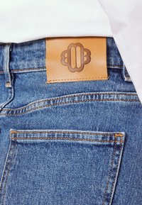 Jean en denim bleu avec un patch de marque en cuir marron visible et une partie d’une chemise blanche rentrée dans la ceinture.