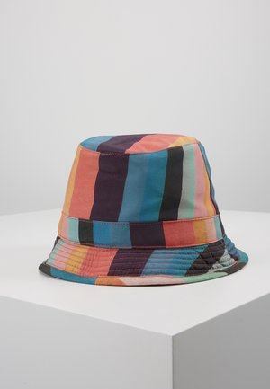 Kleurrijke bucket hat met verticale strepen in oranje, blauw, zwart, roze en geel, geplaatst op een wit voetstuk tegen een effen grijze achtergrond.