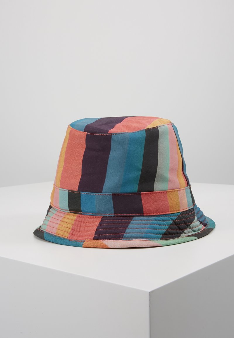Chapeau bob coloré avec des rayures verticales orange, bleu, noir, rose et jaune, posé sur un socle blanc sur fond gris uni.