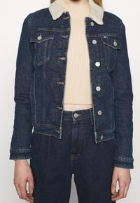 Woman wearing dark blue denim jacket with white shearling collar, beige ribbed cropped sweater, and matching dark blue denim jeans.