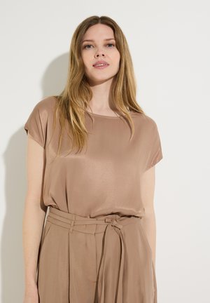 Frau mit langen blonden Haaren, die eine lockere beige Seidenbluse und hoch taillierte Hosen mit einem gebundenen Gürtel trägt, steht vor einem hellen Hintergrund.