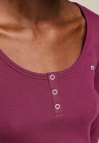 Rippenschaliger burgunderfarbener Oberteil mit Rundhalsausschnitt, drei metallischen Knopfakzenten und einem kleinen Logo in der Nähe der Schulter.