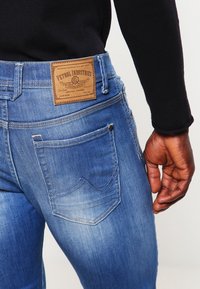 Blå denimjeans med en lädermärke, slim fit, mid-rise, synlig sömnad och två bakfickor. Slät textur och blekt utseende.