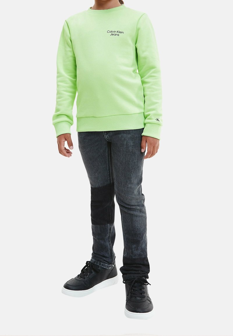 Jasně zelená mikina s žebrovanými manžetami, doplněná tmavými džínami s černou nášivkou a černými teniskami s bílými podrážkami.