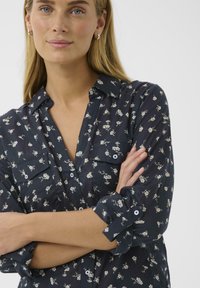 Femme à la peau claire et aux cheveux blonds portant des boucles d'oreilles en argent, une chemise boutonnée à fleurs bleu marine avec les manches retroussées, les bras croisés.