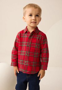 Camicia a quadri rossa con bottoni, dettagli in verde e giallo, tasca sul petto e maniche lunghe. Abbinata a pantaloni di denim blu scuro.