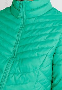 Veste matelassée turquoise avec fermeture éclair à l'avant et col montant, montrant le haut du torse et la zone des épaules.