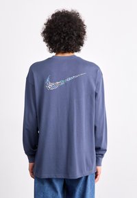 Hosszú ujjú, sötétkék póló puha anyagból, színes Nike logóval a hátoldalán és laza szabással. Farmernadrág látható.