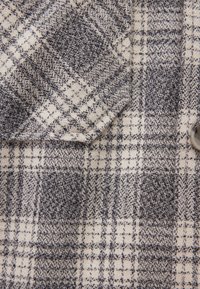 Grau- und cremefarbenes Karostoff mit weicher Textur, der ein gewebtes Muster und eine Knopfdetails aufweist, das unterschiedliche Farbtöne und Gitterlinien hervorhebt.