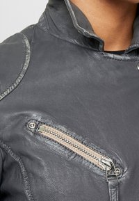 Nærbillede af en mørkegrå læderjakke, der viser en skrå frontlynlås-lomme og en del af den høje krave nær nakken.