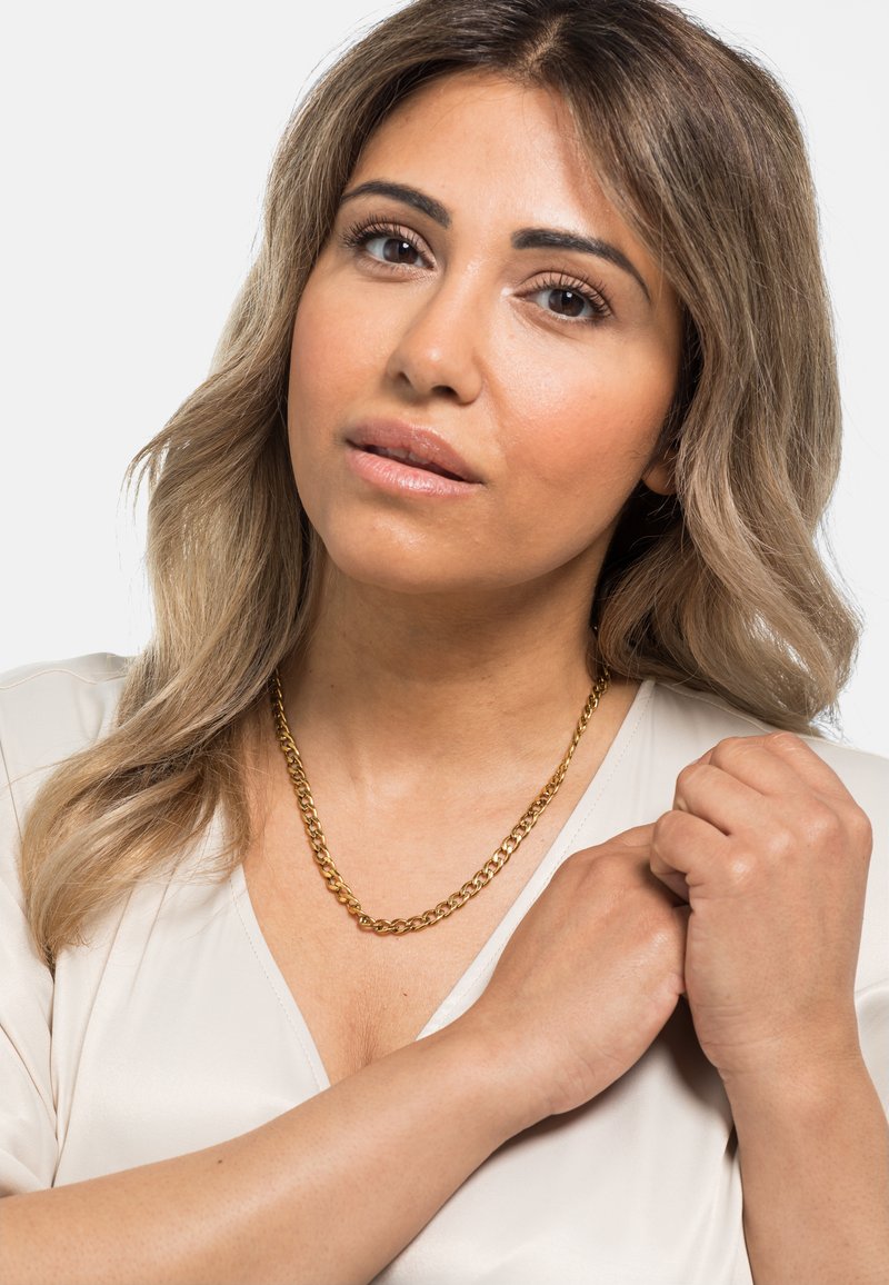 Goldene Kette mit verflochtenen Gliedern, über einem hellen Oberteil getragen. Das Model hat schulterlanges, lockiges Haar und hält ihre Hände in der Nähe des Halsbereichs.