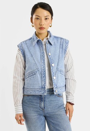 Mujer asiática que lleva un chaleco vaquero sin mangas de color azul claro sobre una camisa blanca con rayas y botones y unos vaqueros azules de cintura alta, de pie de frente.