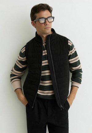 Mężczyzna w czarnej pikowanej kamizelce na beżowo-zielonej pasiastym, długim rękawie, czarnych spodniach i okularach, stojący przy białej ścianie.