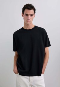 Tricou negru din bumbac cu gât rotund, mânecă scurtă și croială lejeră. Poartă-l cu pantaloni albi, pe un fundal neutru.