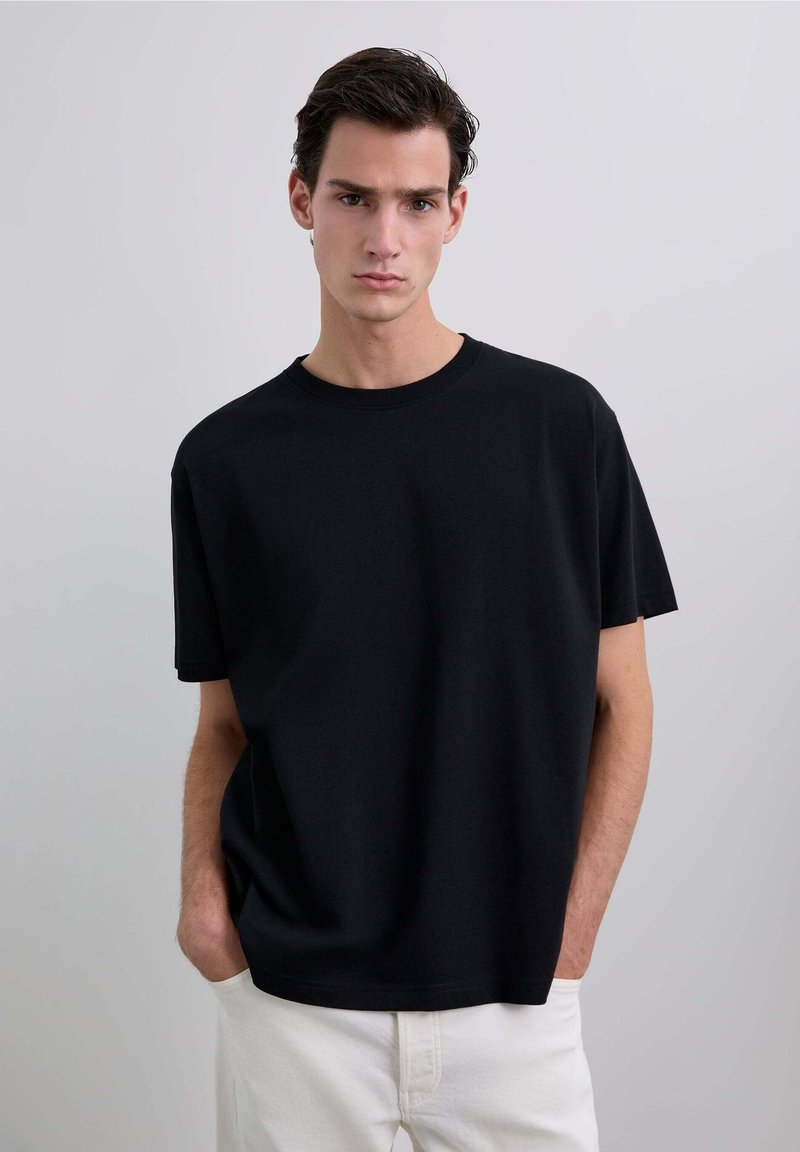 Tricou negru din bumbac cu gât rotund, mânecă scurtă și croială lejeră. Poartă-l cu pantaloni albi, pe un fundal neutru.