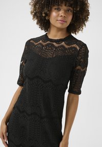 Vestido de encaje negro con mangas cortas, patrones intrincados, un escote con forma de ondas y un cuerpo texturizado.