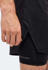 Κοντινό πλάνο ενός χεριού δίπλα σε μαύρα αθλητικά σορτς με το λογότυπο "Peak Performance", εν μέρει καλυμμένα από μαύρο πουκάμισο με σχισμή στο τελείωμα.