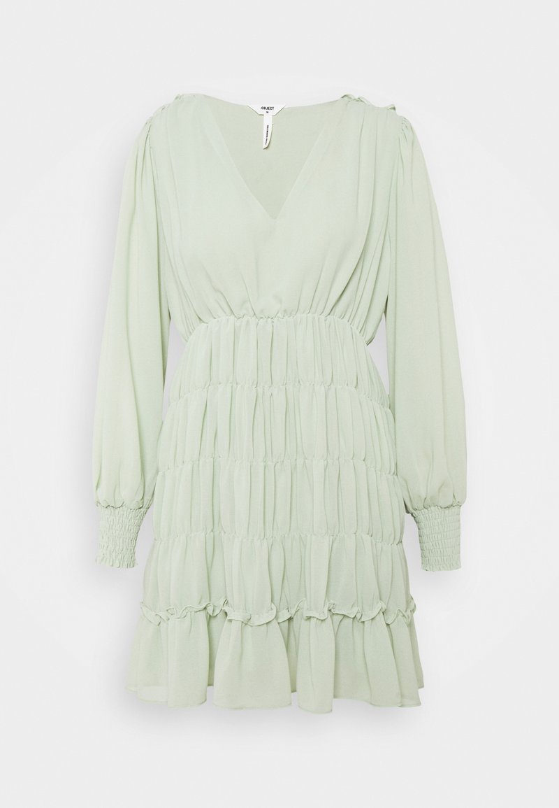 Vaaleanvihreä mekko, joka on valmistettu pehmeästä kankaasta, jossa on V-pääntie, pitkät puffihihat, rypistytty vyötärö ja kerroksellinen röyhelöhelma.