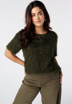 Vrouw met lang donker haar, gekleed in een donkergroene pluizige korte mouwen top en olijfgroene broek tegen een eenvoudige lichte achtergrond.