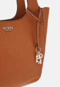 Braune Handtasche aus strukturiertem Leder von Guess mit goldenem Logo und schlüsselähnlichen goldenen Anhängern am Riemen.