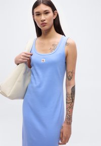 Φόρεμα γαλάζιο με ριχτές ραφές, λαιμόκοψη σε σχήμα scoop και σήμα με λογότυπο. Το μοντέλο κρατά μπεζ τσάντα tote. Τατουάζ φαίνονται στο χέρι.