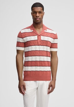 Mann trägt ein kurzärmeliges, gestricktes Poloshirt mit rostfarbenen und weißen Streifen sowie geometrischen Mustern und weiße Hosen, steht vor einem schlichten Hintergrund.