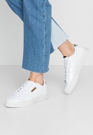 Baskets blanches en cuir lisse, avec des détails perforés et un accent doré au niveau du talon. Portées avec un jean bleu clair effiloché.