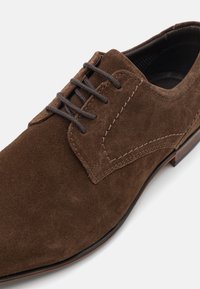 Chaussure habillée en suede marron avec un bout arrondi, dotée de lacets marron foncé, d'un design piqué et d'une semelle extérieure marron contrastante.