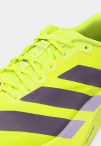 Heller gelber Laufschuh mit lila Streifen, mit einem Mesh-Obermaterial, strukturierter Oberfläche und einem weißen Logo auf der Zunge.