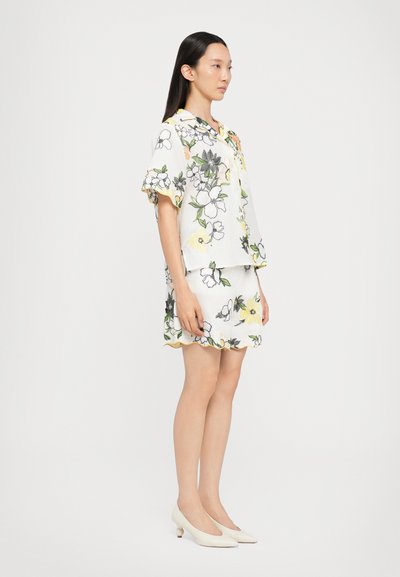 Blomstrede bluse og shorts sæt i hvid, med grønne, gule og sorte accenter. Bølgekant og korte ærmer. Hvide sko.