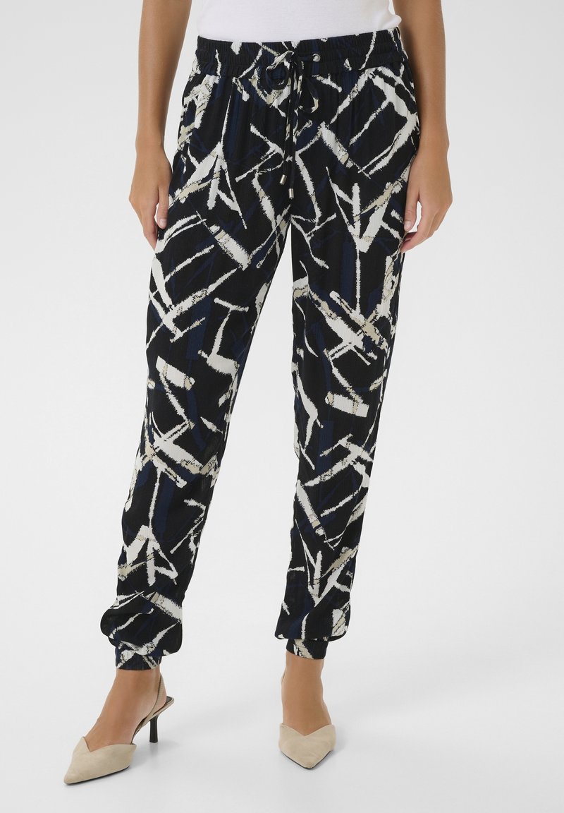 Femme portant un pantalon à imprimé abstrait noir et blanc avec une taille élastique et un cordon de serrage, associé à des escarpins beiges à bout pointu.