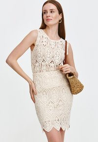 Sukienka koronkowa w kolorze kremowym z kwiatowymi wycięciami, dopasowanym gorsetem i falbaniastym dołem, modelka trzymająca plecioną torebkę.
