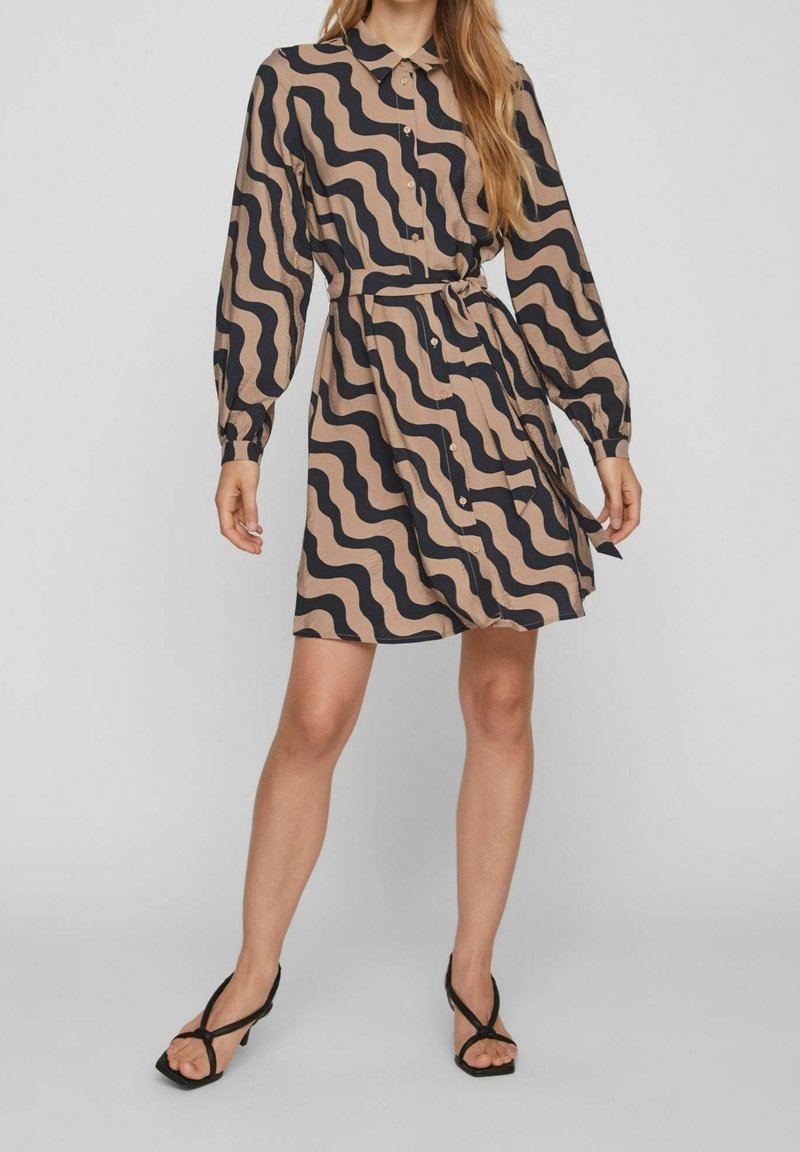 Femme portant une robe longue jusqu'aux genoux à manches longues avec des rayures ondulées noires et beiges et des talons ouverts noirs, debout devant un fond uni.