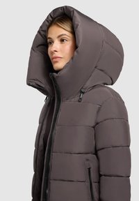 Pufasta jakna u prigušenoj smeđoj boji s velikim, strukturalnim kapuljačom, debelim štitnim dizajnom i prednjim patentnim zatvaračem. Ima bočne džepove na zip.