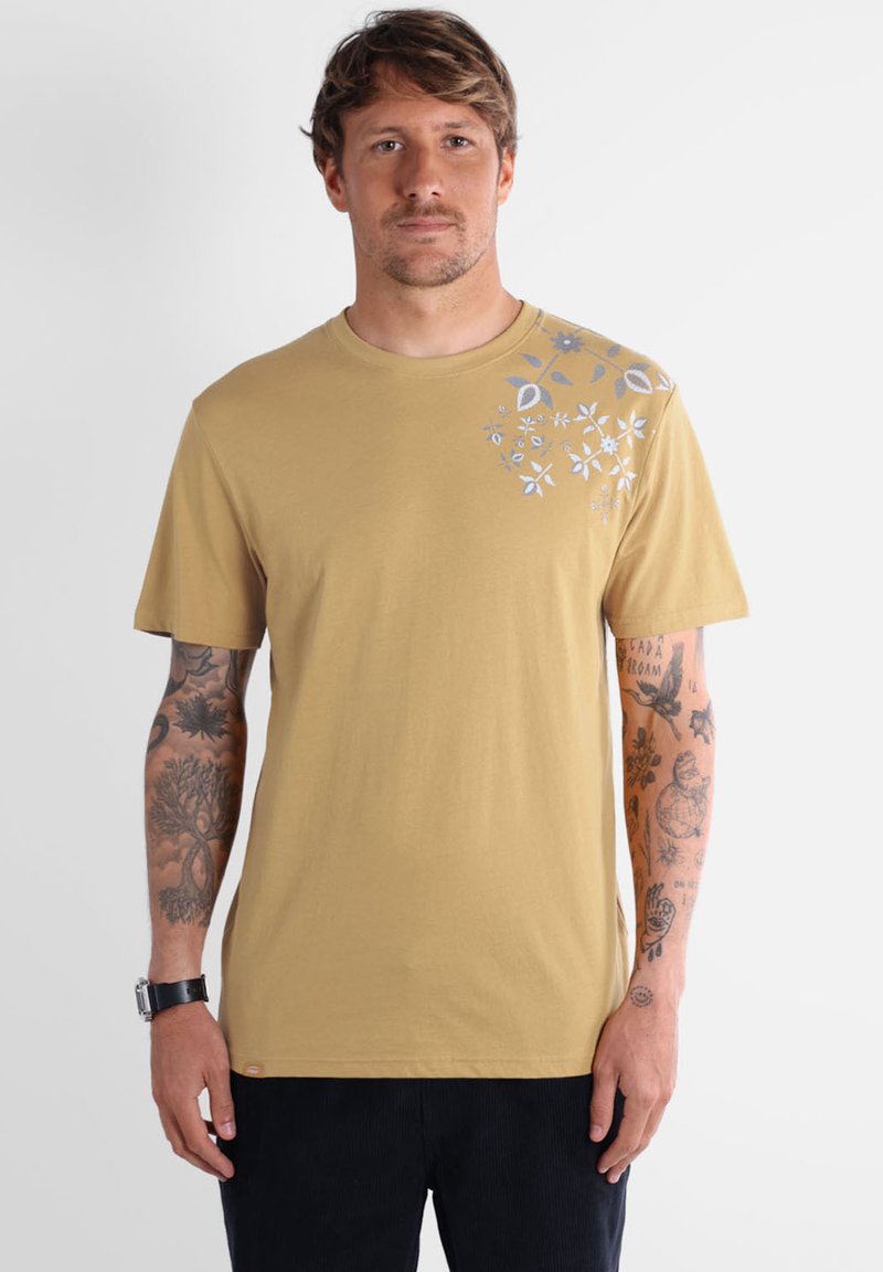 T-shirt en coton beige avec un col rond et des manches courtes, décoré de motifs floraux bleus sur l'épaule gauche.