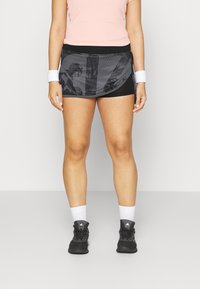 Svart träningskjol med ett genomskinligt, mönstrat lager ovanpå, ihopparat med en ljusrosa topp och svarta sneakers. Vit handledsband och strumpor synliga.