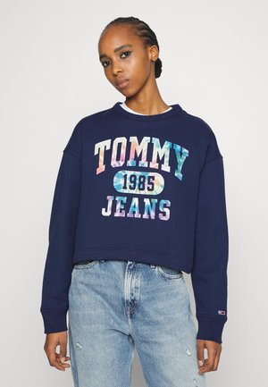 Granatowa krótka bluza z kolorowym napisem "TOMMY JEANS 1985", posiadająca okrągły dekolt i długie rękawy. Noszona z jasnymi dżinsami.