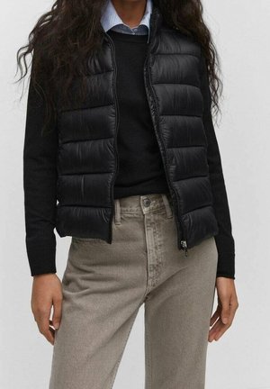Mujer con chaleco acolchado negro sobre un jersey negro y vaqueros beige claro, con cabello largo, oscuro y rizado visible.