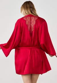 Accappatoio in raso rosso con pannello posteriore in pizzo, caratterizzato da maniche ampie, vita con cintura e una superficie liscia e riflettente.