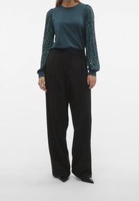 Top blu petrolio a maniche lunghe con maniche a sbuffo decorate di paillettes e pantaloni neri a vita alta. I pantaloni hanno un taglio sartoriale e pieghe sottili.