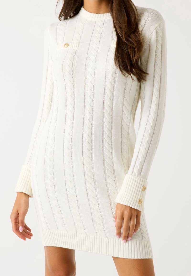 Femme portant une robe-pull en maille torsadée blanche avec manches longues, poignets côtelés avec boutons, et un petit détail de poche poitrine.