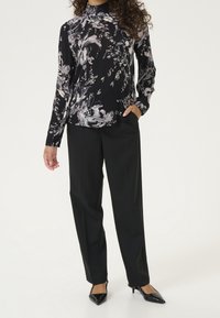 Blouse noire à motif floral avec un col haut froncé, associée à un pantalon noir ajusté et des escarpins noirs à bout pointu.