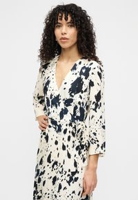 Φόρεμα τύπου wrap σε κρεμ χρώμα με έντονο μαύρο μοτίβο πιτσιλισμάτων, βαθύ V λαιμόκοψη και μακριά μανίκια. Δένει στη μέση για ρυθμιζόμενη εφαρμογή.