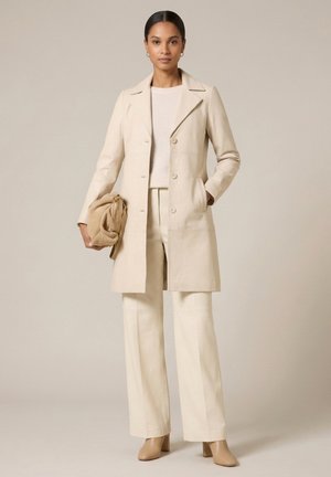 Kvinde iført beige lang frakke, vidde bukser, let sweater og brune ankelsstøvler, der holder en sammenfoldet brun håndtaske mod en beige baggrund.